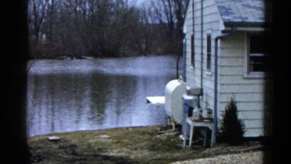 The camera shifts to the side of the house, showing windows, yard objects, and calm lake water behind the property.