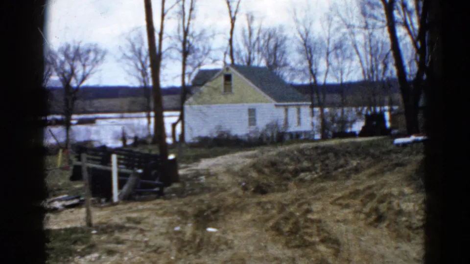 A closer portion of the pan shows the white house and yellow gable beside water, with muddy ruts and debris in the foreground.