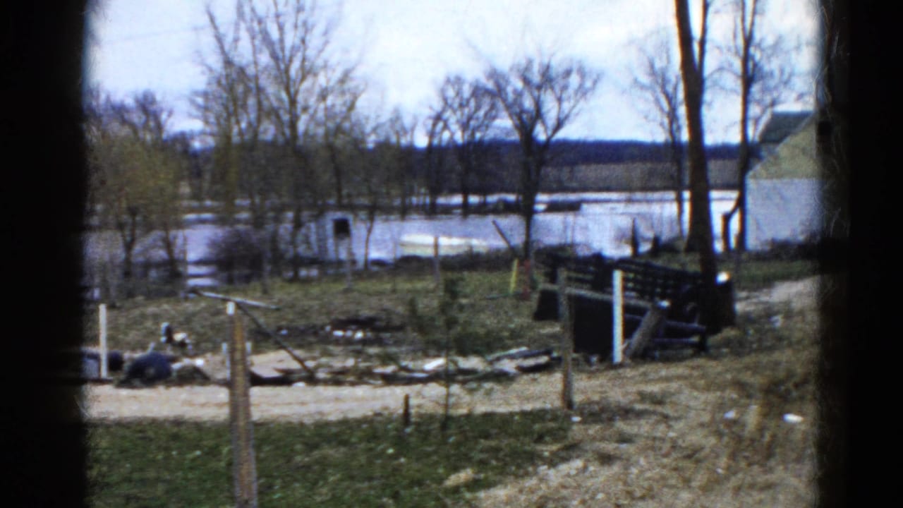 Flooded rural field with bare trees, fencing, debris, and a distant light-colored house.