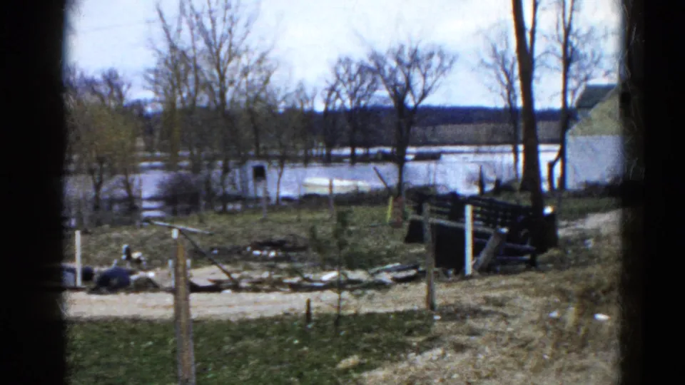 1961 Wauconda, Illinois 8mm frame showing floodwater beyond bare trees, with fence posts, debris, grass, and a distant house.