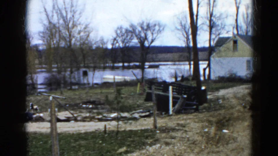 1961 Wauconda, Illinois home-movie frame of a flooded field seen past fence rails and bare trees, with a house on the right.
