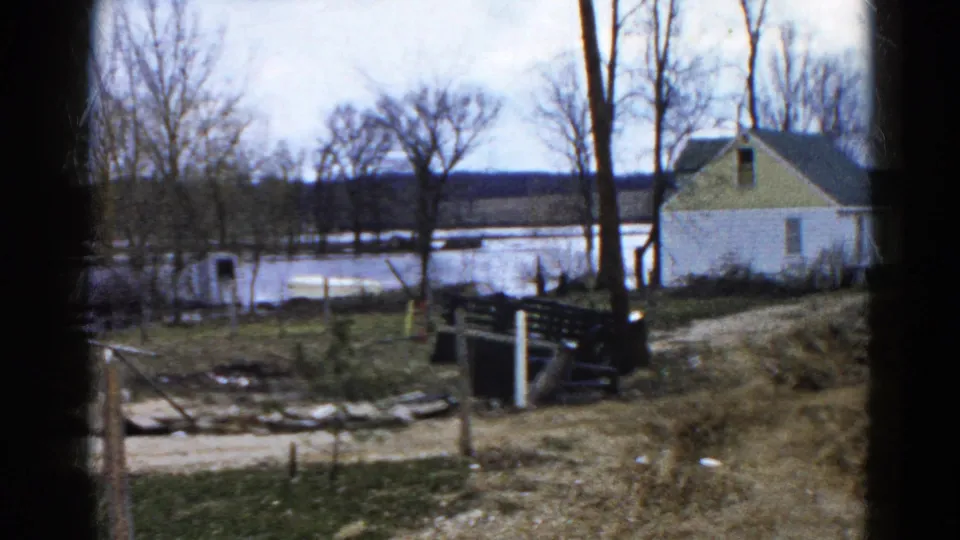 1961 Wauconda, Illinois 8mm frame showing standing water behind leafless trees, a house, fence posts, and uneven rural ground.