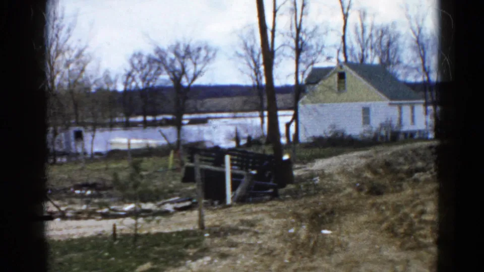 1961 Wauconda, Illinois frame showing a flooded rural area beside a dirt path, with bare trees, fence rails, debris, and a house.