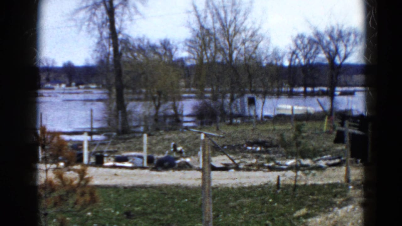 Flooded riverbank seen past a dirt road, fence posts, bare trees, and scattered debris.