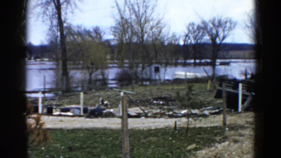 1961 Wauconda view of a flooded riverbank with a dirt road in front, bare trees behind, fence posts, debris, and a small white structure.