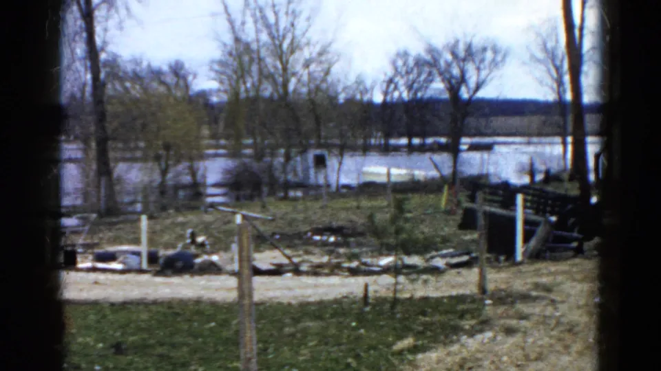 1961 Wauconda frame showing floodwater, a dirt road, leafless trees, muddy grass, scattered debris, posts, and a dark bench or railing.