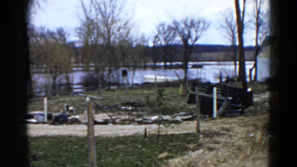 1961 Wauconda 8mm frame of a flooded riverside area, with a dirt road, bare trees, fence posts, debris, grass, and a dark bench or railing.
