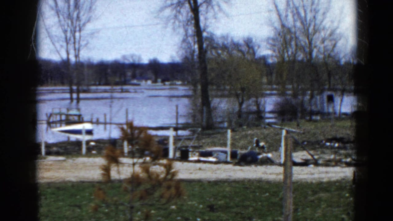 Flooded lakeside area seen through a dark window frame with docks, boats, trees, and shoreline debris.