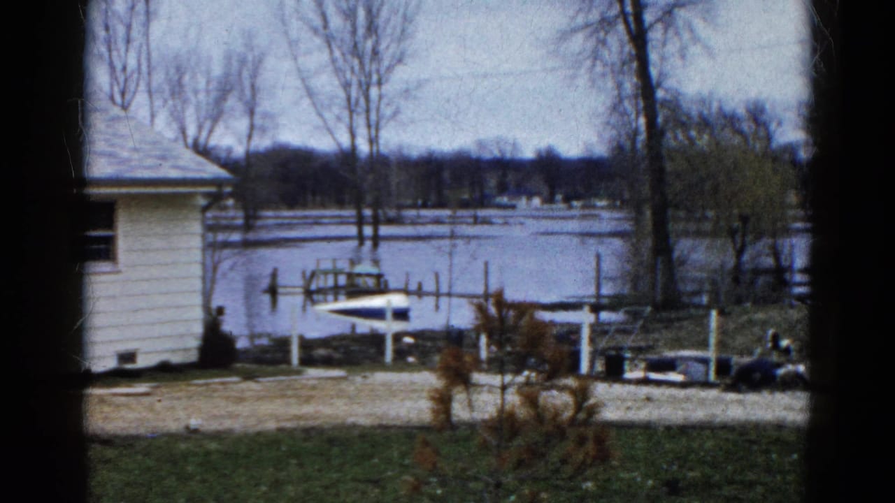 Winter lakeshore with white house, dock posts, small boat, bare trees, and icy water.