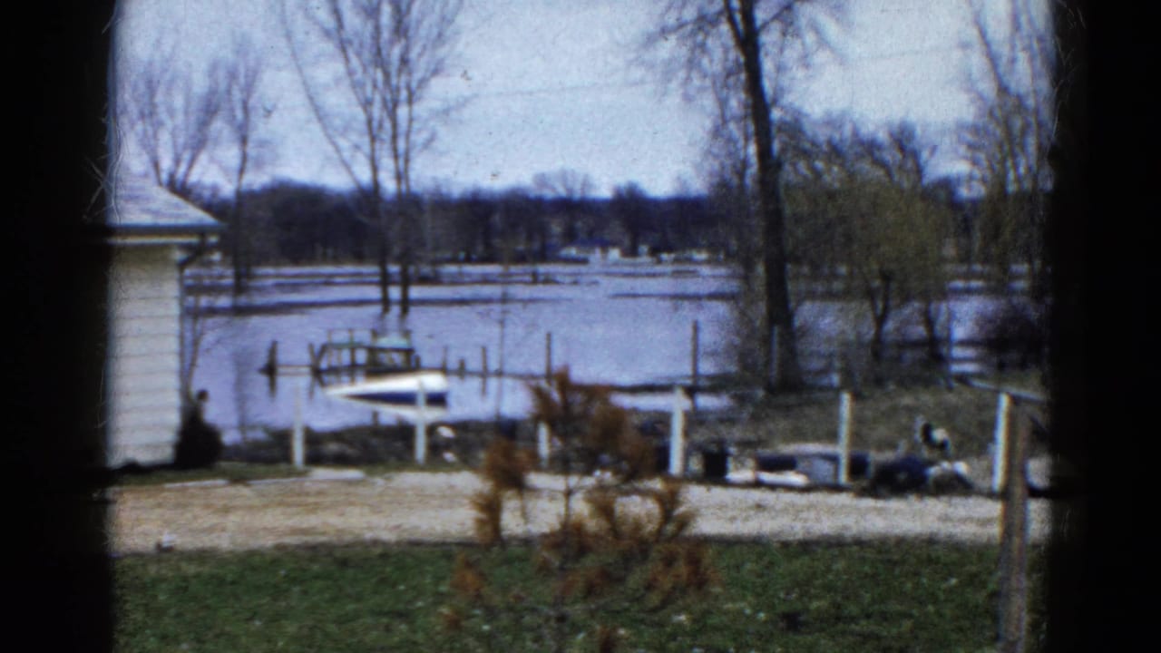 Small boat near flooded dock posts with a house edge, bare trees, and icy lake water.