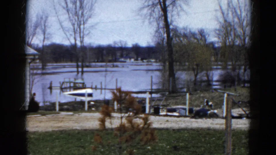 1961 Wauconda, Illinois home-movie frame of a winter lakeside yard facing a flooded dock, small boat, bare trees, and icy lake.