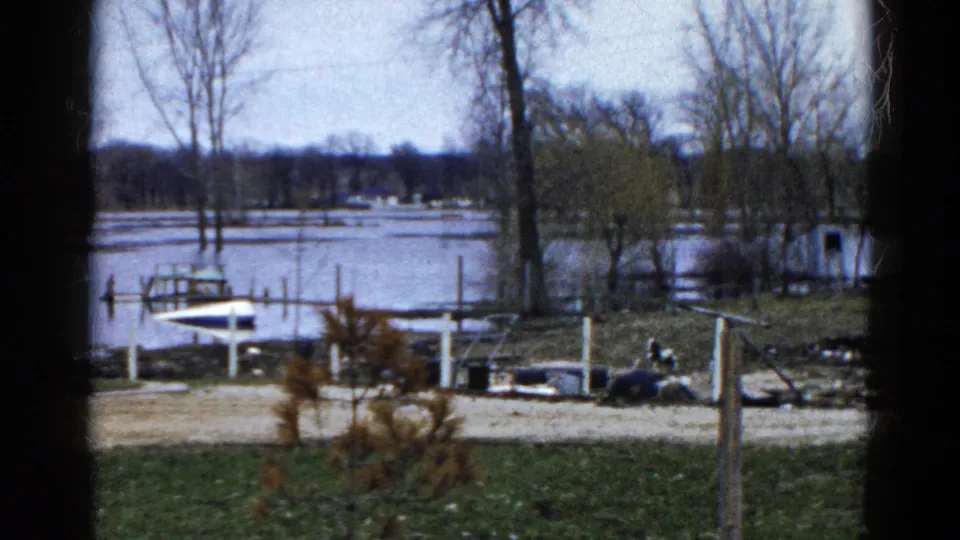1961 Wauconda lakeshore frame showing flooded dock posts, a small boat, bare trees, winter grass, icy water, and distant homes.