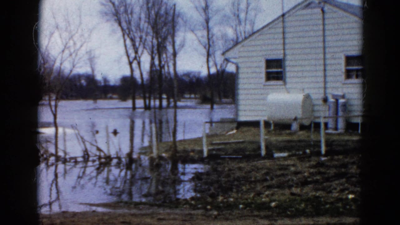 Floodwater surrounds a white clapboard house with bare trees and a propane tank nearby.