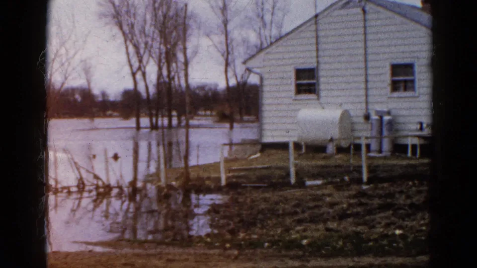 1961 Wauconda, Illinois frame with a white clapboard house, flooded yard, bare trees, reflected water, and utility equipment near the wall.