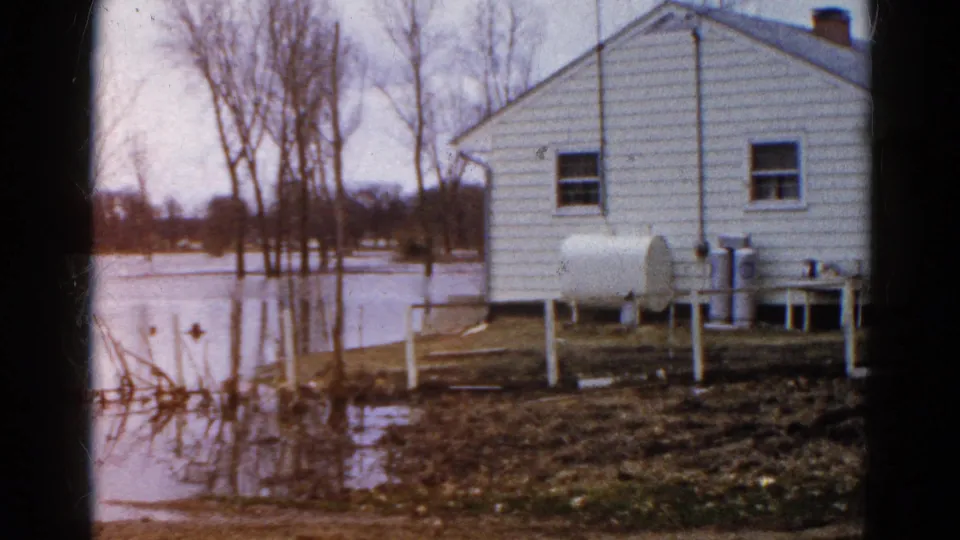 1961 Wauconda, Illinois home-movie frame showing a flooded residential yard, white house wall, propane tank, bare trees, and muddy ground.
