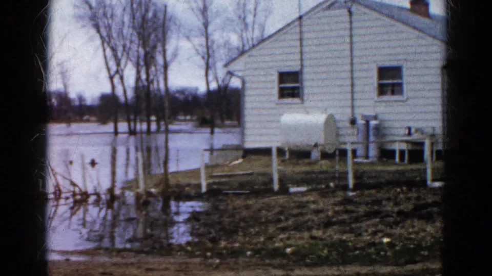 1961 Wauconda, Illinois frame showing the side of a white clapboard house on raised supports, floodwater, bare trees, and a propane tank.