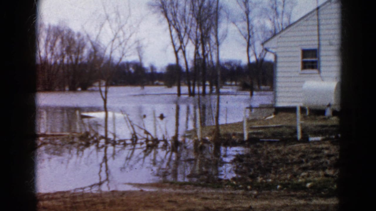 Floodwater surrounds a white house, bare trees, fence posts, and a propane tank.