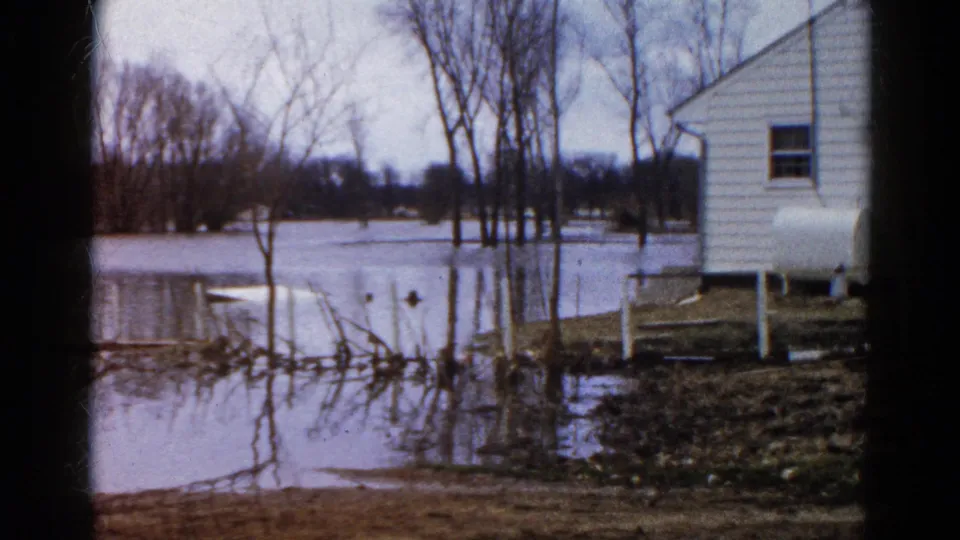 1961 Wauconda, Illinois home-movie frame showing floodwater around a white house, bare trees, fence posts, and a raised propane tank.