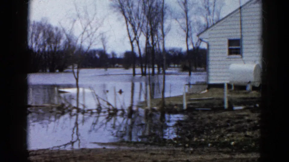 1961 Wauconda, Illinois frame with a white clapboard house, standing floodwater, bare trees, and a propane tank close to the waterline.