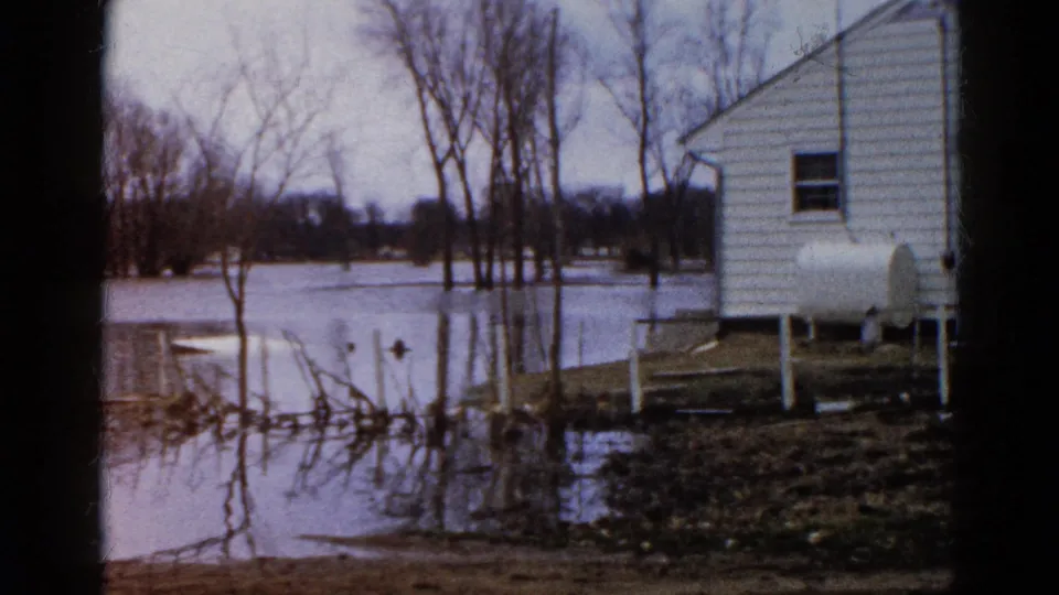 1961 Wauconda, Illinois home-movie view of a flooded yard, white house, raised propane tank, bare trees, and branch reflections in still water.