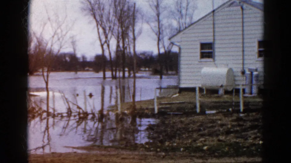 1961 Wauconda, Illinois frame showing a white residential building, propane tank, bare trees, muddy ground, and floodwater across the property.