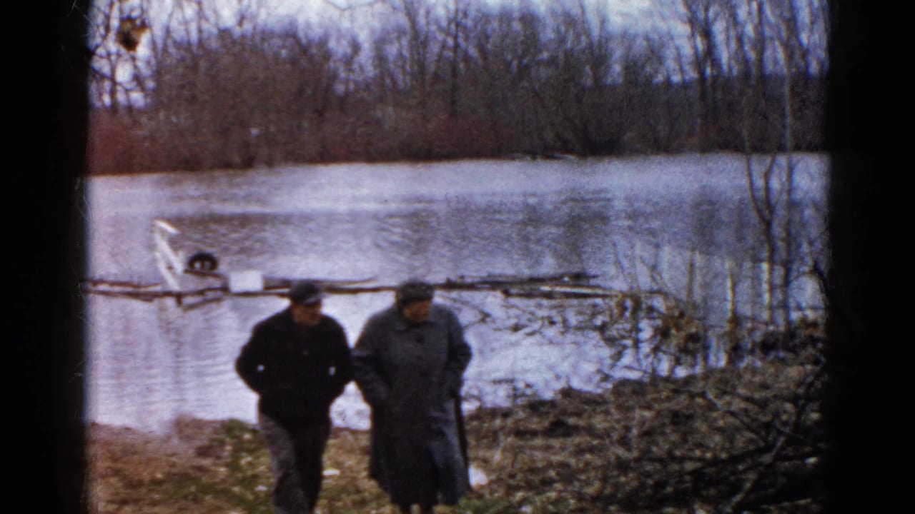 Two bundled adults walk beside floodwater near bare trees and muddy ground.