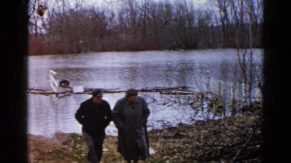 In 1961 Wauconda, Illinois footage, two adults in heavy coats walk along a muddy bank beside high water and leafless trees.