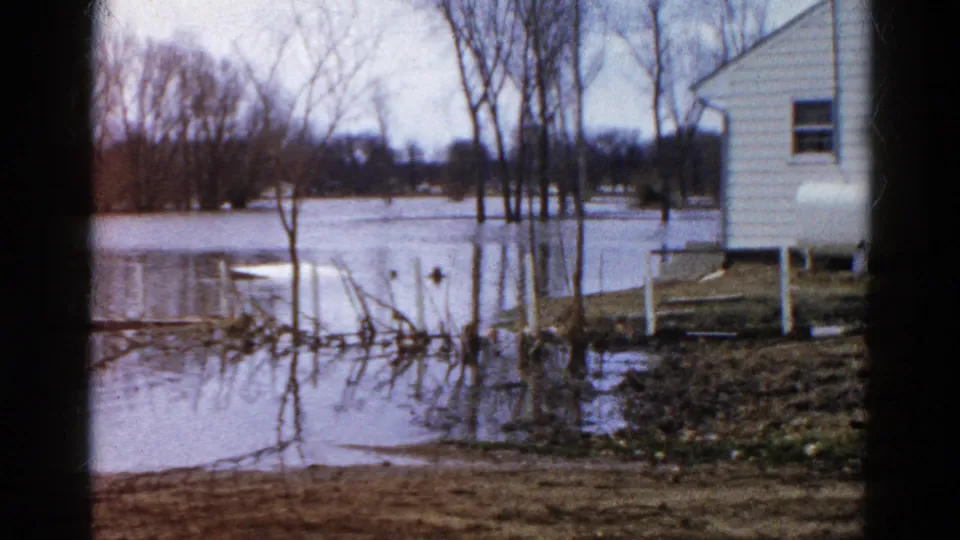 High water stands among bare trees and near a white house in Wauconda, Illinois, with muddy ground and submerged posts visible.