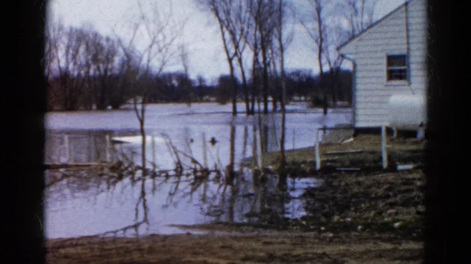 The 1961 home-movie frame shows a flooded Wauconda yard, bare trees reflected in water, and the side of a white house.