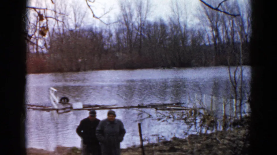 Two men in heavy coats walk along the muddy Wauconda shoreline, with floodwater, bare trees, and a small boat visible in the background.