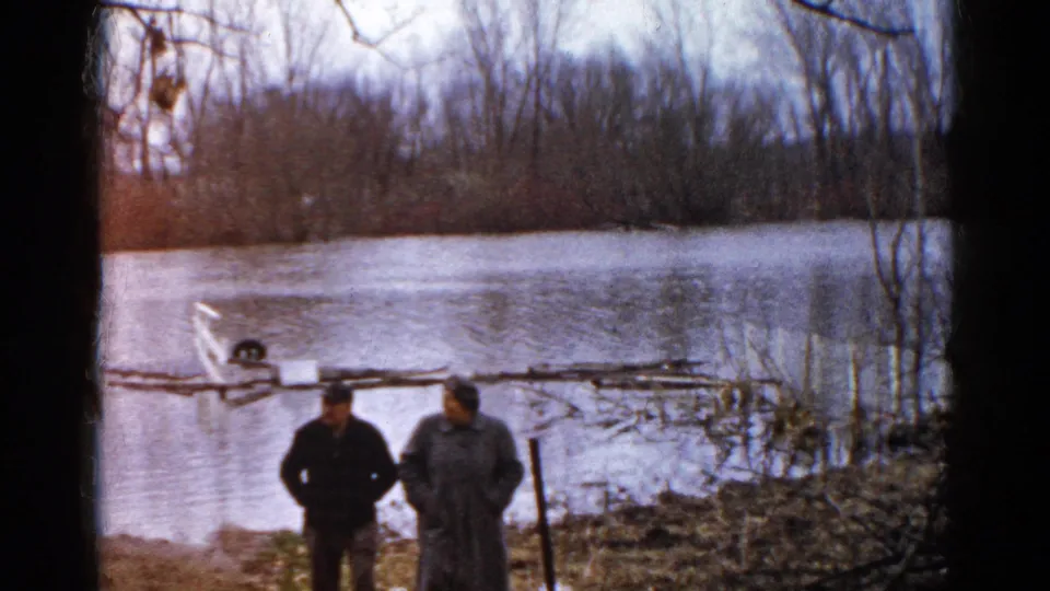 The home-movie frame shows two men beside a flooded Wauconda waterway, with a small boat, brush, and winter-bare trees across the scene.