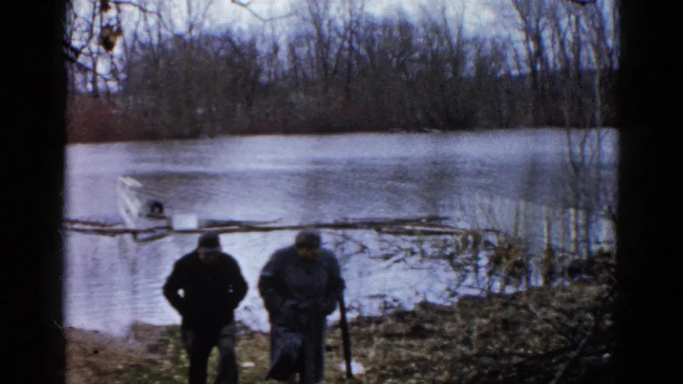 At the end of the 1961 Wauconda clip, two men face downward near the muddy flooded bank, with a partly submerged boat and tree-lined water behind them.