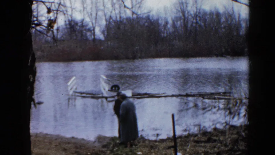 In supplied 1961 Wauconda, Illinois context, two bundled adults stand by a flooded pond with bare trees and a partly submerged object nearby.
