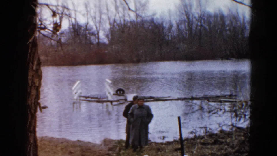 In this 8mm home-movie frame, two adults stand on the bank of a flooded pond in the supplied Wauconda, Illinois location.
