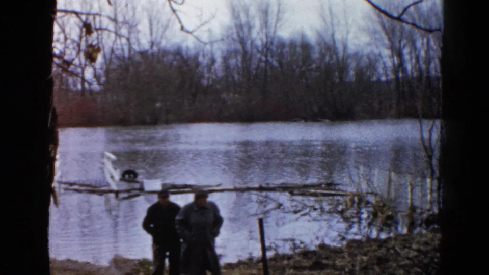 Two adults stand side by side at the flooded pond in the supplied 1961 Wauconda, Illinois home-movie footage, with bare trees behind them.