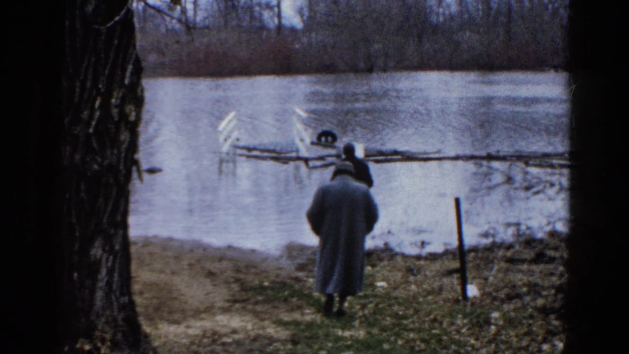 Two adult men by a Wauconda lake in 1961, one in a long coat and hat, framed by dark tree trunks.