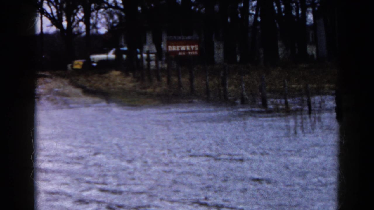 Floodwater covers a rural road with bare trees, fence posts, a white vehicle, and a red sign in the distance.
