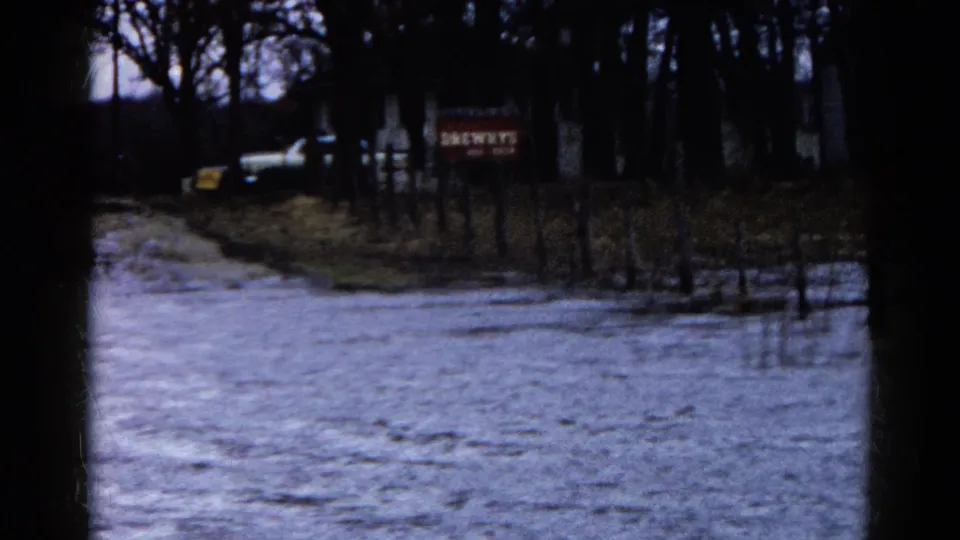 1961 Wauconda frame of floodwater across a rural road, with dark tree trunks, fence posts, a white vehicle, and a red Drewry's sign.