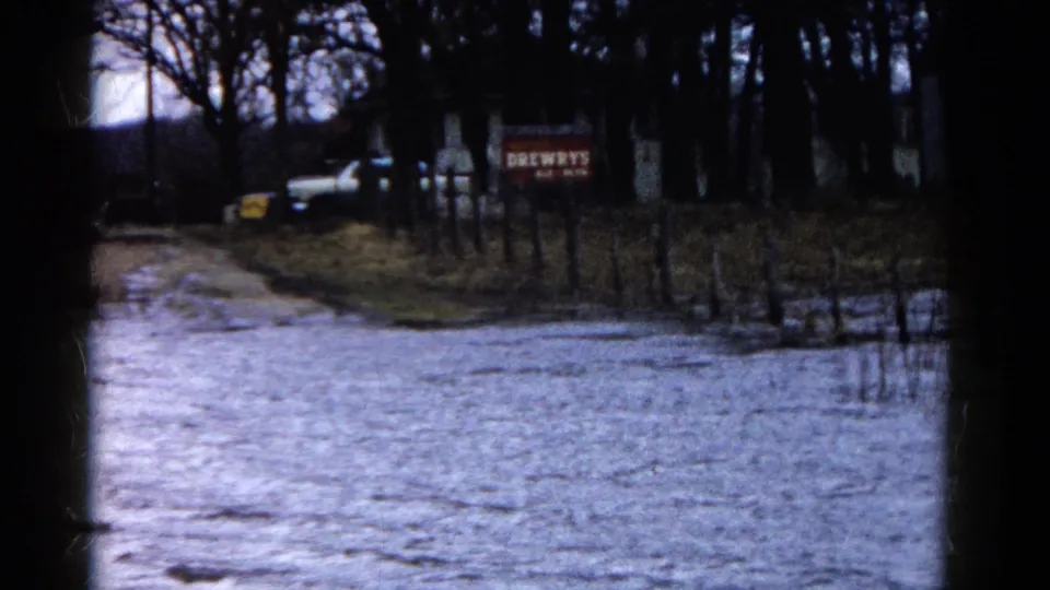 1961 Wauconda home-movie view of a water-covered road, roadside fencing, bare trees, a distant white vehicle, and Drewry's signage.
