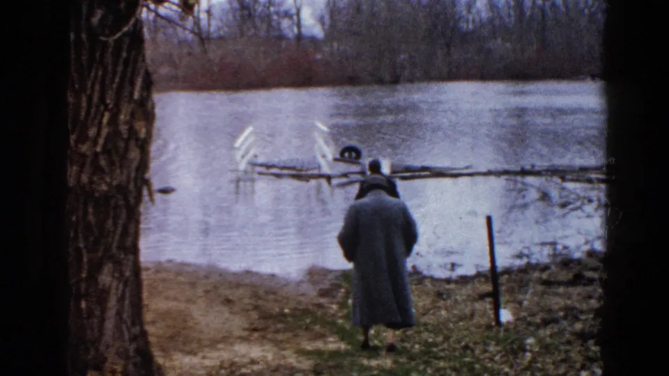 1961 Wauconda riverbank frame showing a person in a long coat watching floodwater around a partly submerged white structure.