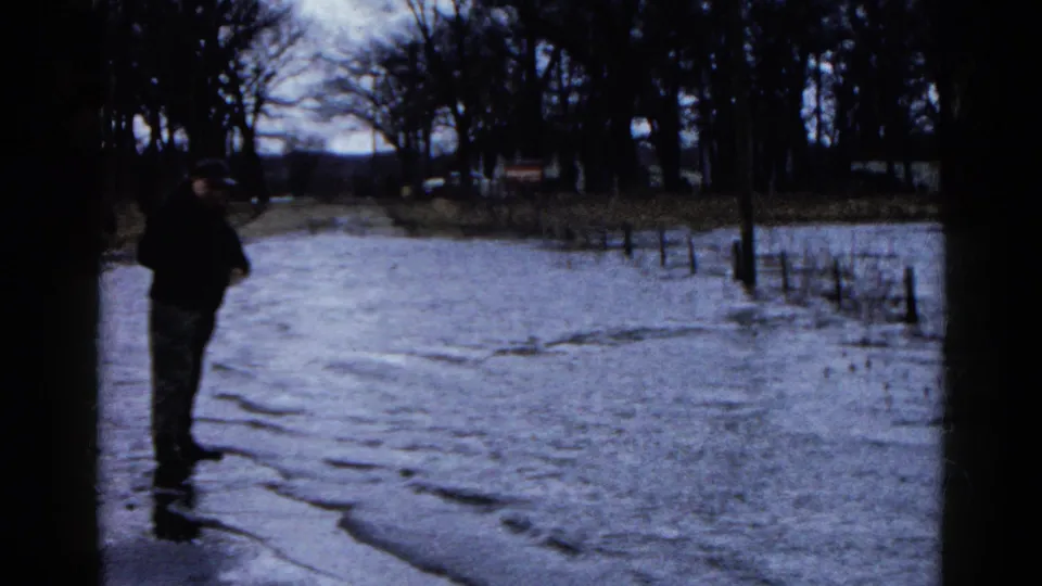 In Wauconda, Illinois, a man appears to fish beside a flooded road lined with bare trees in 1961 home-movie footage.