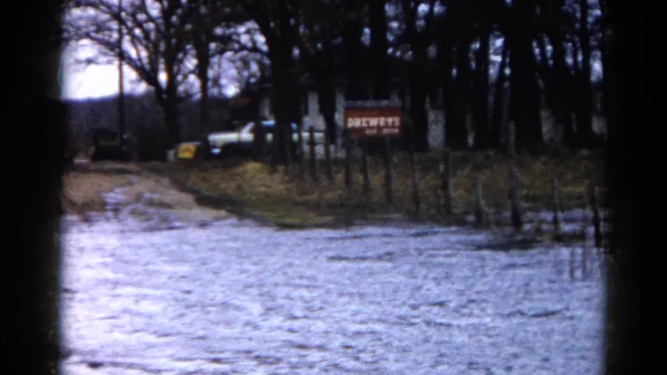 A wide 1961 Wauconda home-movie frame shows floodwater on a muddy roadside with fencing, trees, a parked vehicle, and a BREWERS sign.