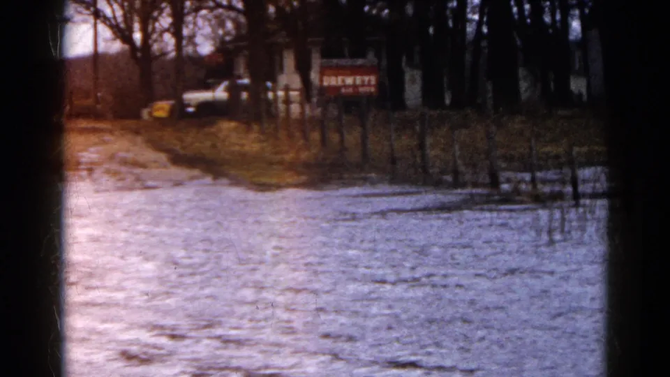 The flooded roadside in Wauconda, Illinois, curves past fence posts toward a house or outbuilding, parked vehicle, and BREWERS sign.