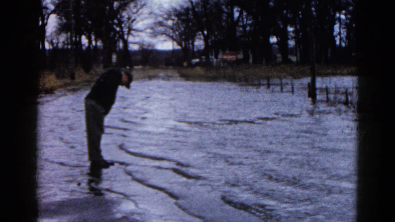In Wauconda, Illinois, a man in dark clothing leans toward floodwater covering a rural road or property in 1961 home-movie footage.