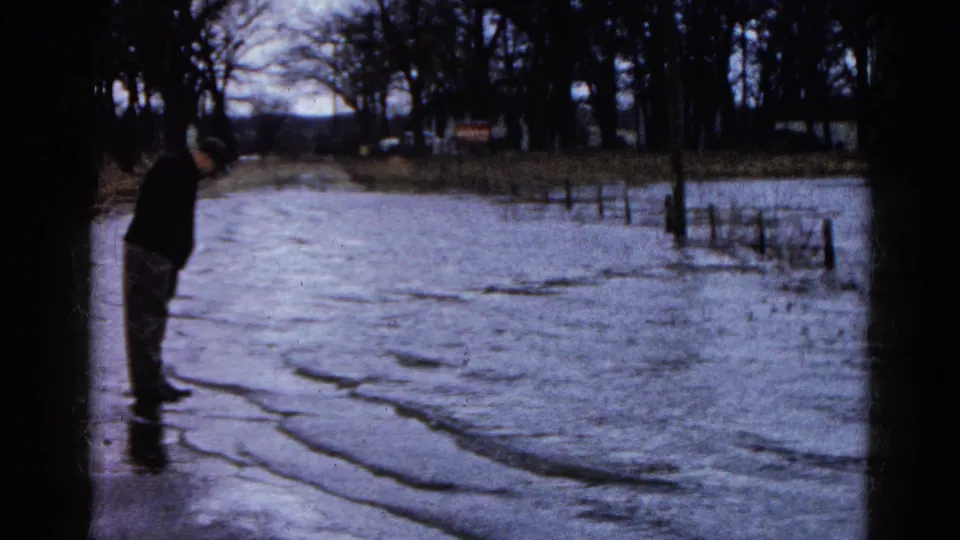The man stands beside floodwater in Wauconda, Illinois, with bare trees, fence posts, and distant vehicles visible in the 1961 frame.