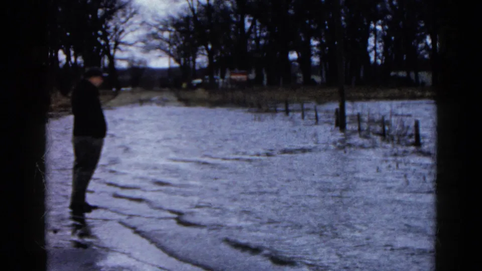 At the end of the clip, the man stands near floodwater in Wauconda, Illinois, with submerged fencing and bare trees beyond him.