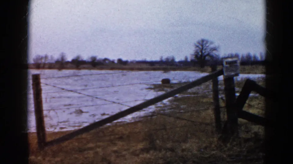 1961 Wauconda, Illinois home-movie frame of a wet or icy field bordered by a wooden gate, fence posts, bare trees, and open winter sky.