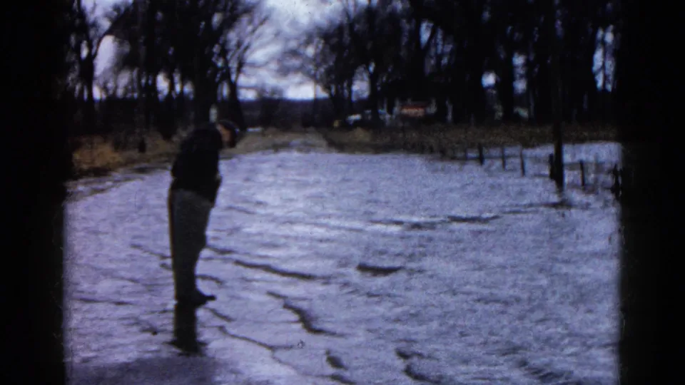 1961 Wauconda, Illinois frame showing an adult man in a dark jacket and light pants standing in shallow floodwater near leafless trees.