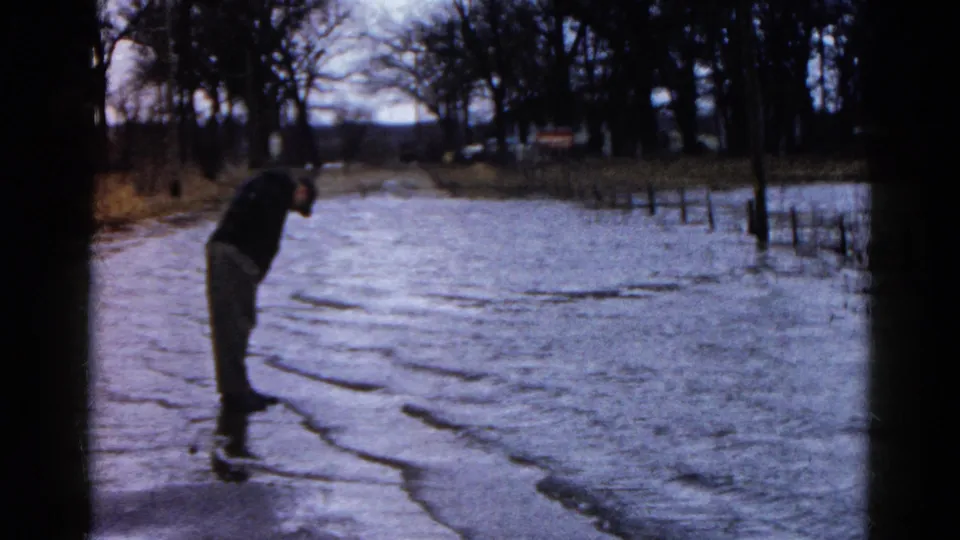 1961 Wauconda, Illinois home-movie frame of an adult man bending to inspect his feet while standing in cold flooded ground near trees and fencing.