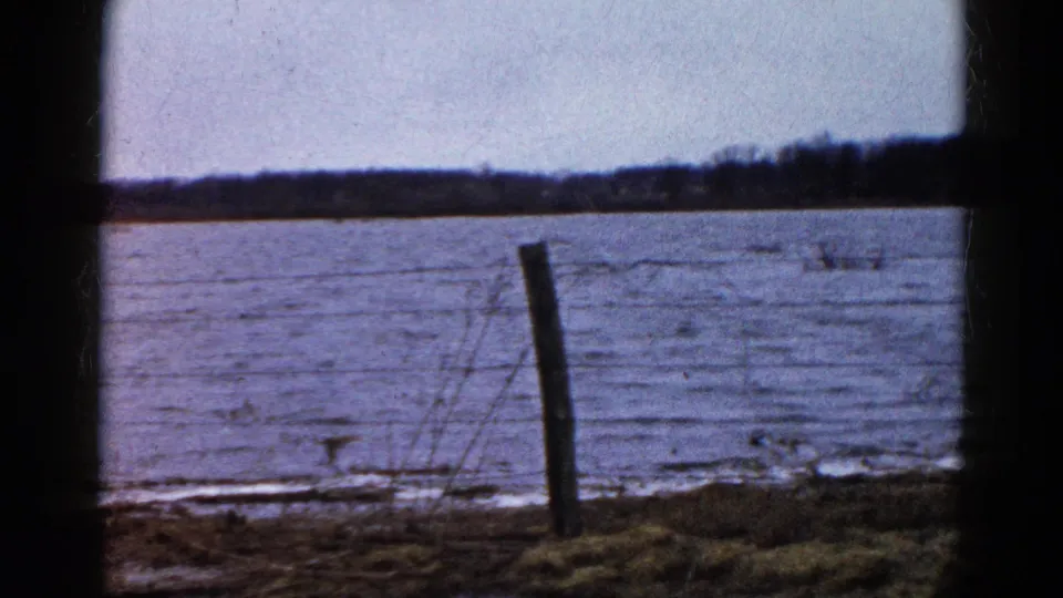 1961 Wauconda, Illinois home-movie frame showing floodwater beyond a leaning fence post and barbed wire, with trees on the horizon.